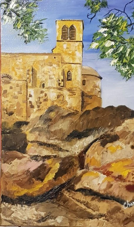 Église en pierre surplombant des rochers jaunis, sous un ciel bleu et quelques feuillages.
